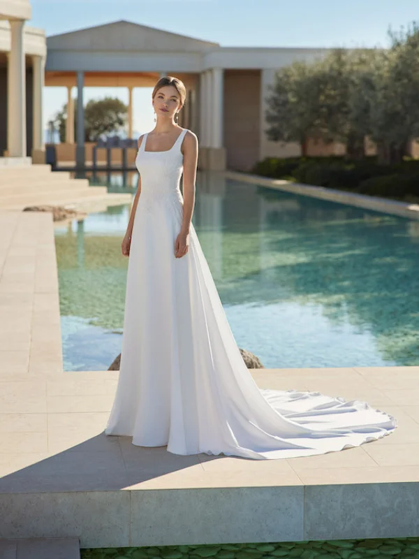 Une mariée portant une robe de mariée blanche avec une coupe droite et un col carré, posant élégamment devant une piscine à l'architecture classique.