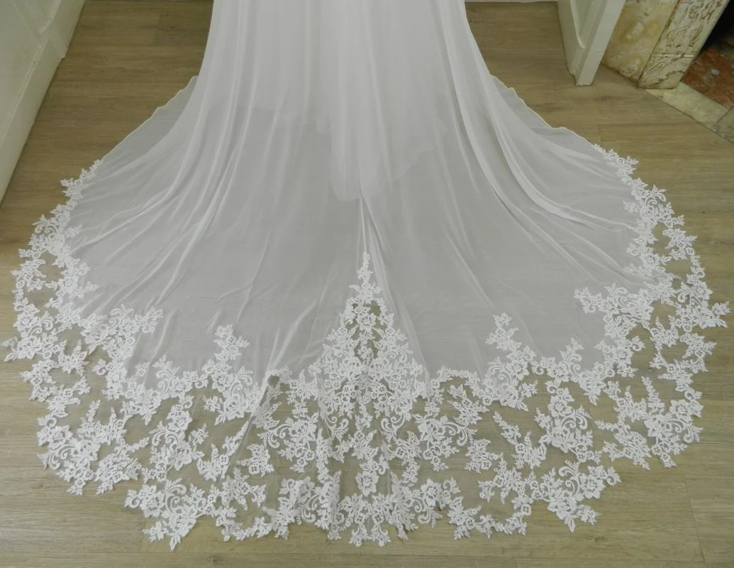 Gros plan technique au sol sur les finitions d'une longue traîne en mousseline ivoire bordée d'une large guipure de dentelle florale festive pour la robe 44275 Justin Alexander.