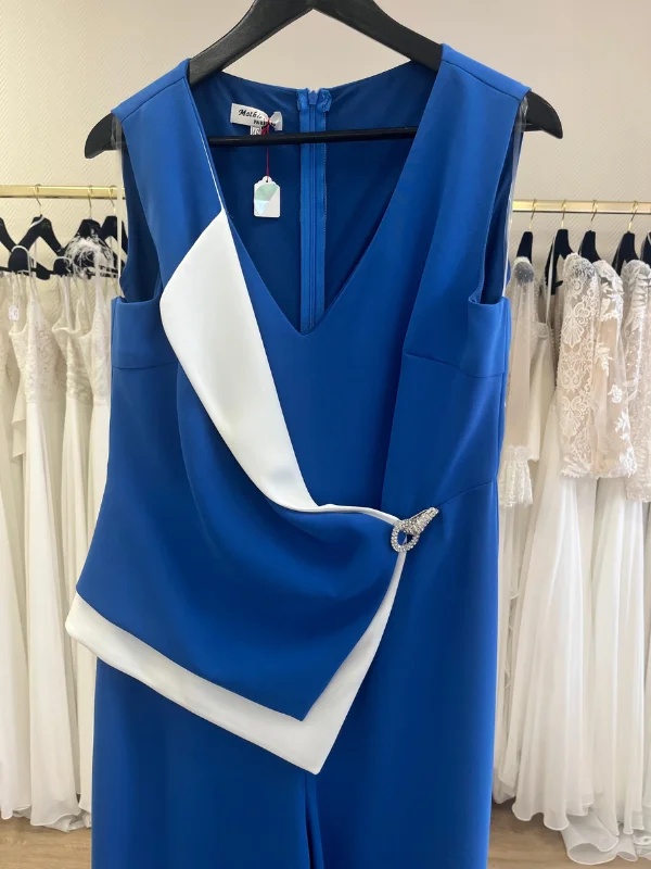 Gros plan du buste de la combinaison bleu royal Laurièle de Mathieu Paris, montrant le décolleté en V, le rabat asymétrique drapé blanc et la boucle strassée à la taille, suspendue sur un cintre en boutique.