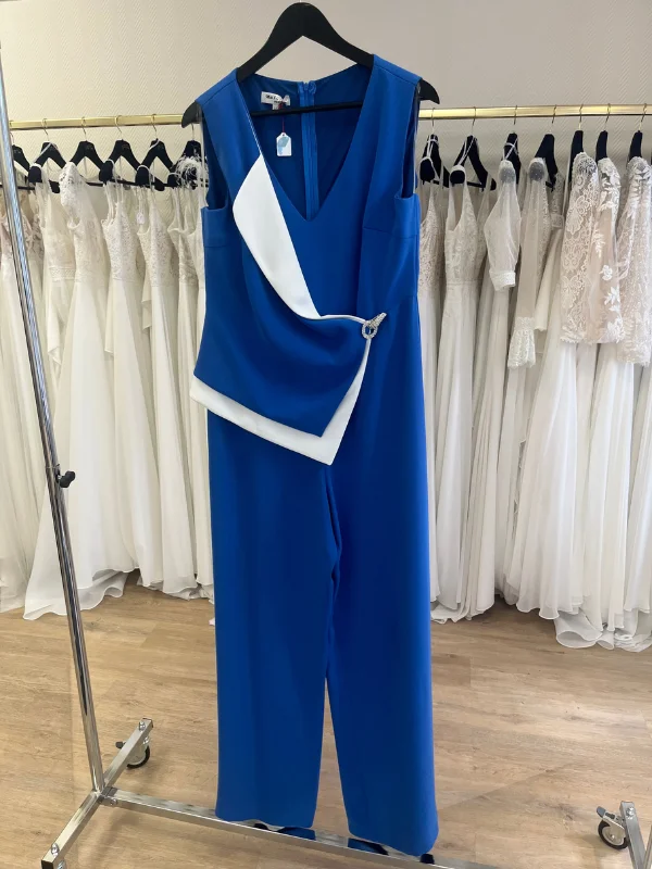 Combinaison bleu royal Laurièle de Mathieu Paris suspendue de face sur un cintre dans la boutique Petite Folie. La tenue présente un col en V, un drapé asymétrique blanc avec une boucle dorée à la taille et des jambes larges, devant un portant de robes de mariée.