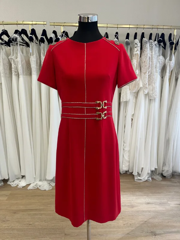 Robe de cocktail rouge Carla de Sonia K présentée de face sur un buste mannequin dans la boutique Petite Folie. La tenue intègre un liseré doré vertical, une double ceinture intégrée à boucles métalliques dorées et des boutons dorés aux épaules.