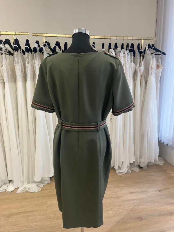 Robe de cocktail kaki Cupidon de Sonia K présentée de dos sur un buste mannequin en boutique. La tenue montre une fermeture éclair discrète au centre du dos, des empattements aux épaules, une ceinture ajustée à galon rayé et une coupe droite ajustée.