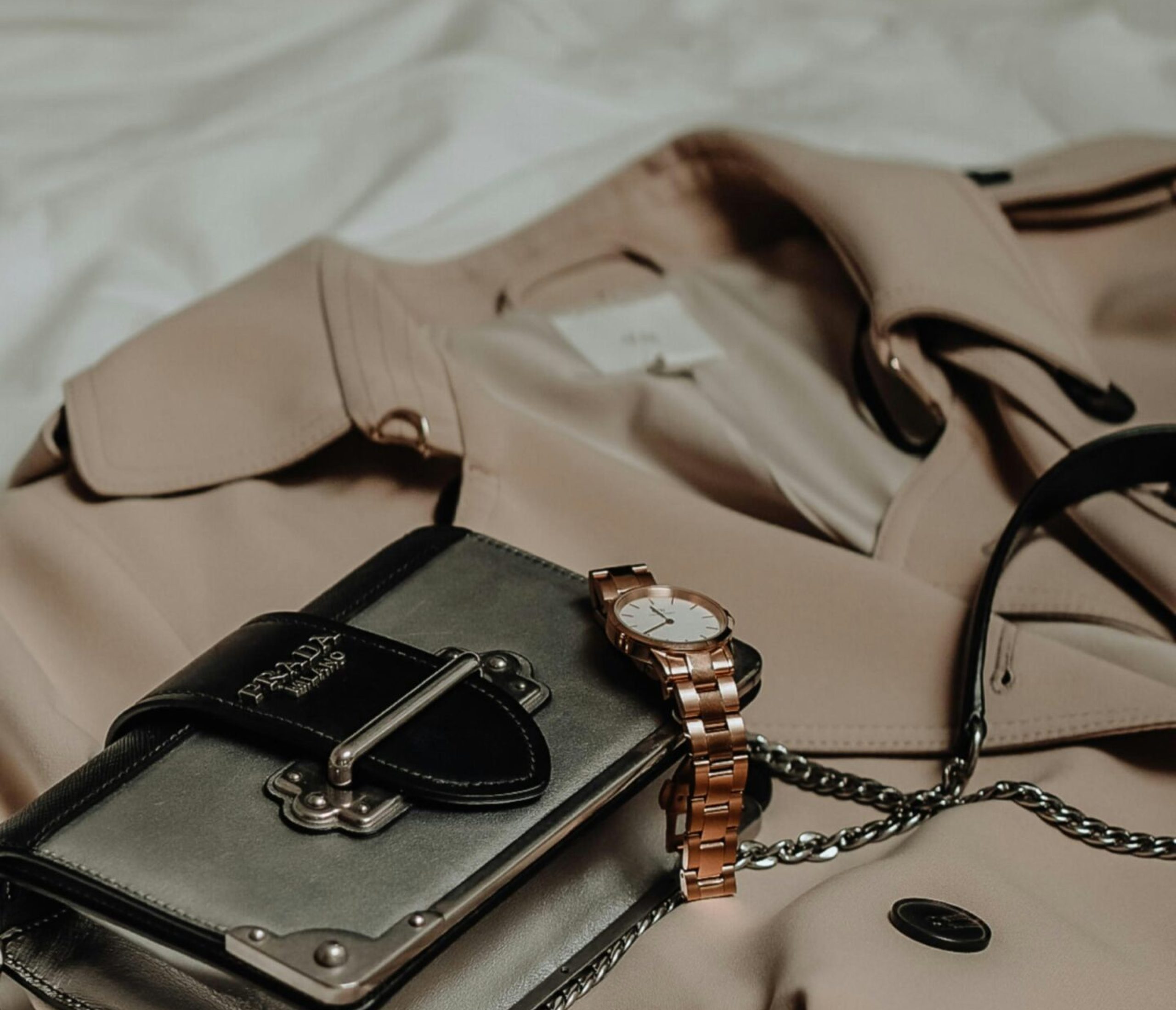 Accessoires Sac à main argenté sur un trench beige – Accessoires de luxe – Petite Folie Accessoires de luxe argenté avec chaîne posé sur un trench beige, disposé sur un lit avec un éclairage doux.