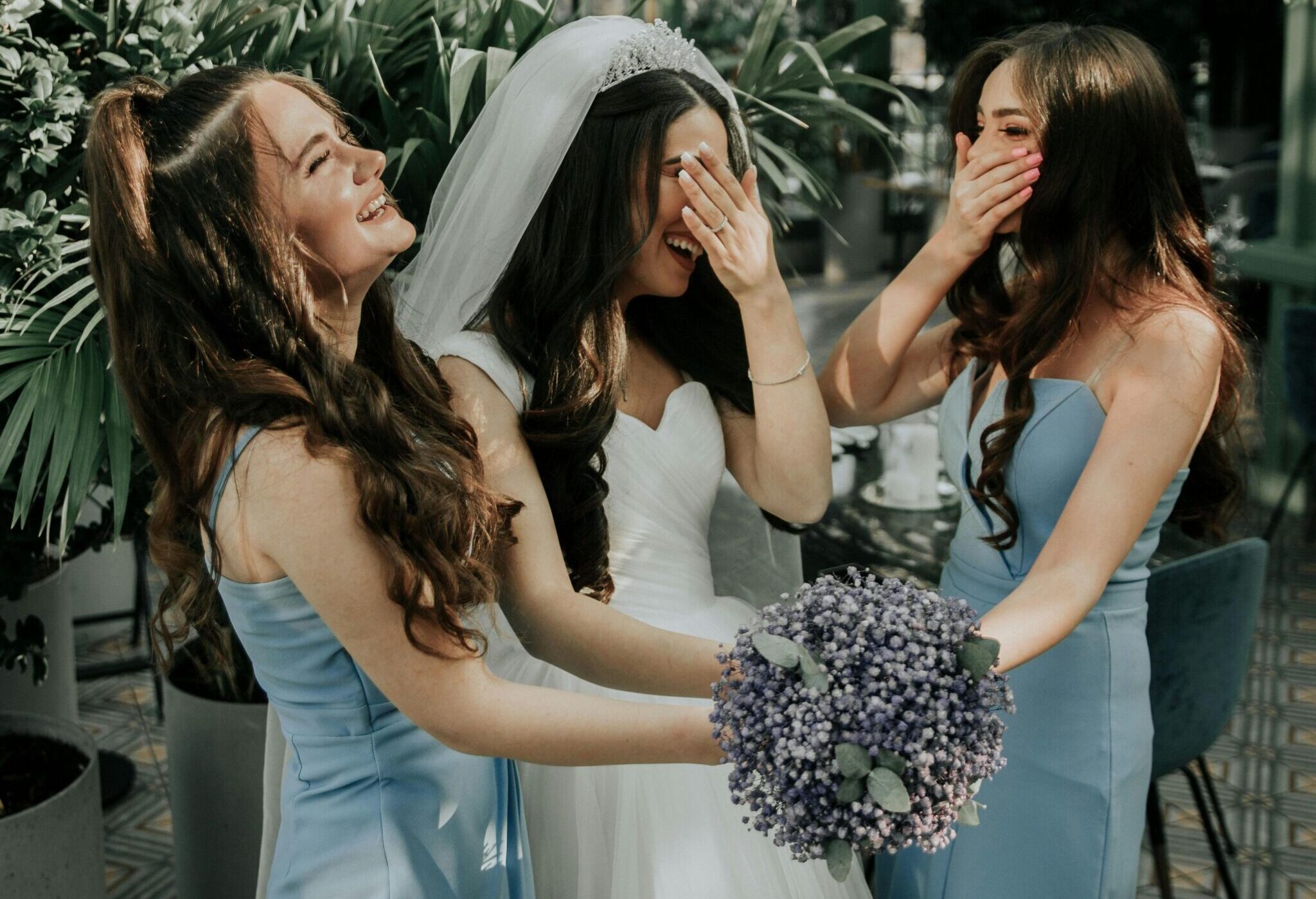 Mariée et demoiselles d’honneur partageant un moment de joie Mariée en robe blanche riant avec ses demoiselles d'honneur en robes bleues, dans un cadre extérieur verdoyant.